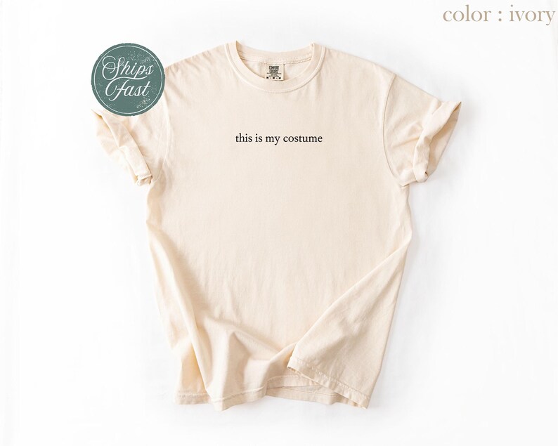 May include: Ivory-colored t-shirt with the phrase "this is my costume" printed in black. The shirt is folded, showing the sleeves and neckline. A round green sticker with the words "Ships Fast" is in the upper left corner.