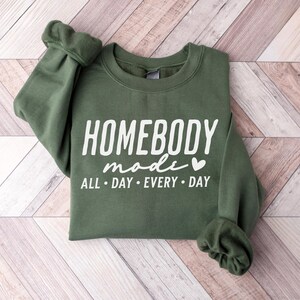Puede incluir: Una sudadera verde con el texto "Homebody mode" y un peque&ntilde;o s&iacute;mbolo de coraz&oacute;n. El texto "All day every day" est&aacute; impreso debajo del texto principal.