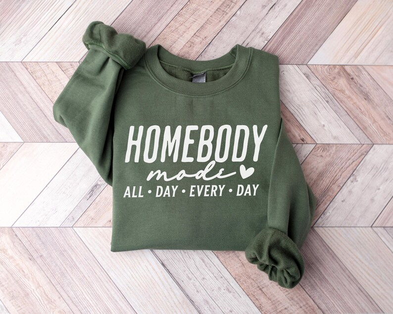 Puede incluir: Una sudadera verde con el texto "Homebody mode" y un peque&ntilde;o s&iacute;mbolo de coraz&oacute;n. El texto "All day every day" est&aacute; impreso debajo del texto principal.