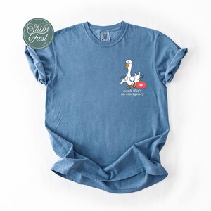 May include: A blue t-shirt featuring a white goose wearing a red first aid kit and the text "honk if it's an emergency".
