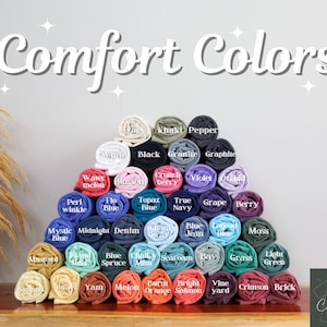 May include: A display of rolled-up t-shirts in various colors, including white, black, and blue. The text "Comfort Colors" is at the top. Each shirt is labeled with its color name, such as "Watermelon" and "Berry".