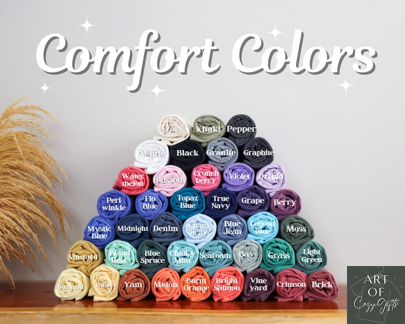 May include: A display of rolled-up t-shirts in various colors, including white, black, and blue. The text "Comfort Colors" is at the top. Each shirt is labeled with its color name, such as "Watermelon" and "Berry".