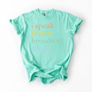 May include: A turquoise t-shirt with the text "i speak fluent broadway" printed on the front in a rainbow of colors.