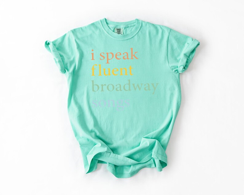 May include: A turquoise t-shirt with the text "i speak fluent broadway" printed on the front in a rainbow of colors.