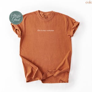 May include: A yam-colored t-shirt with the phrase "this is my costume" printed in white. The shirt has a crew neck and short sleeves, with the sleeves rolled up. A round sticker with the words "Ships Fast" is on the left side.