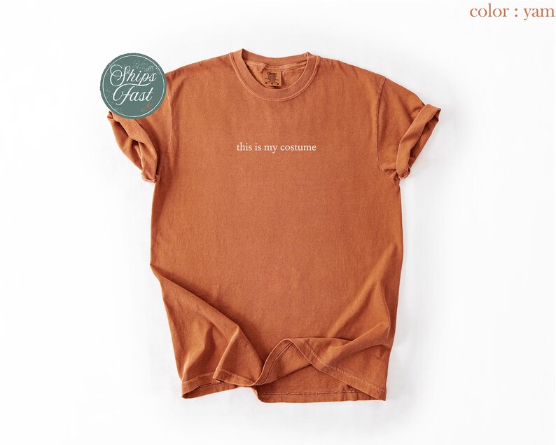 May include: A yam-colored t-shirt with the phrase "this is my costume" printed in white. The shirt has a crew neck and short sleeves, with the sleeves rolled up. A round sticker with the words "Ships Fast" is on the left side.