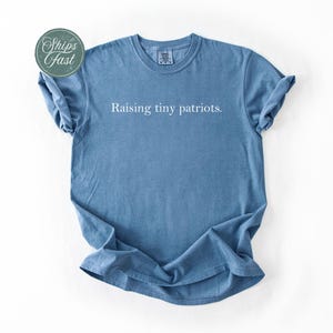 May include: A blue t-shirt with the phrase "Raising tiny patriots" printed in white. The shirt has short sleeves and a crew neck. A green and white sticker that says "Ships Fast" is in the upper left corner.