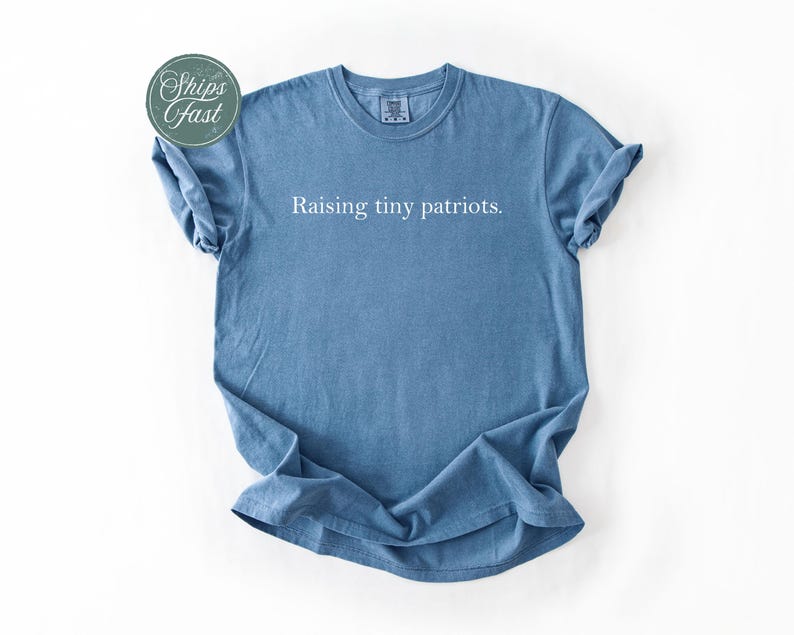 May include: A blue t-shirt with the phrase "Raising tiny patriots" printed in white. The shirt has short sleeves and a crew neck. A green and white sticker that says "Ships Fast" is in the upper left corner.