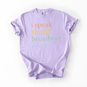 May include: A lavender t-shirt with the text "i speak fluent broadway songs" printed on the front in rainbow colors.
