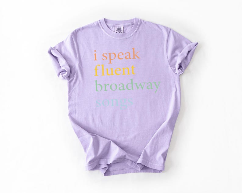 May include: A lavender t-shirt with the text "i speak fluent broadway songs" printed on the front in rainbow colors.