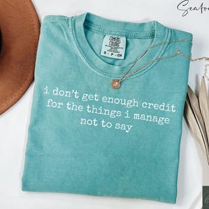 Puede incluir: Camiseta verde mar con el texto blanco "i don't get enough credit for the things i manage not to say". Un collar dorado con un colgante descansa sobre la camiseta. La camiseta tiene una etiqueta Comfort Colors.