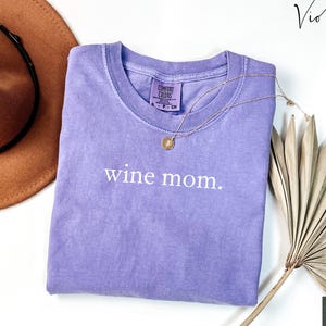 Pu&ograve; includere: Una maglietta viola con la scritta "wine mom." stampata in bianco sul davanti. La maglietta &egrave; piegata e appoggiata su una superficie bianca.