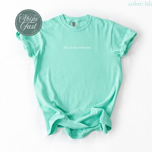 May include: A light turquoise t-shirt with the phrase "this is my costume" printed in white. The shirt has rolled-up sleeves and a crew neck. A circular sticker with the words "Ships Fast" is in the upper left corner.