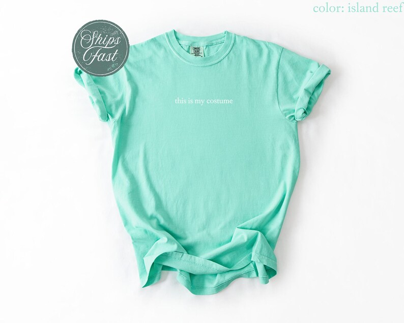 May include: A light turquoise t-shirt with the phrase "this is my costume" printed in white. The shirt has rolled-up sleeves and a crew neck. A circular sticker with the words "Ships Fast" is in the upper left corner.
