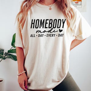 Puede incluir: Una camiseta de color crema con el texto "HOMEBODY mode" y un s&iacute;mbolo de coraz&oacute;n. El texto "ALL DAY EVERY DAY" est&aacute; debajo del texto principal.