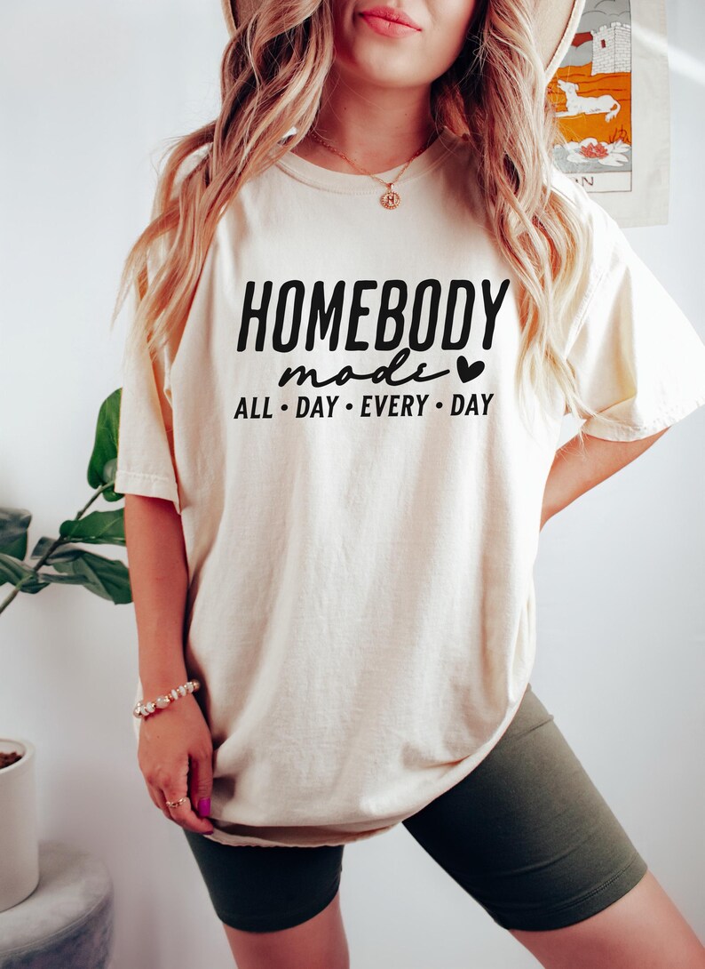 Puede incluir: Una camiseta de color crema con el texto "HOMEBODY mode" y un s&iacute;mbolo de coraz&oacute;n. El texto "ALL DAY EVERY DAY" est&aacute; debajo del texto principal.