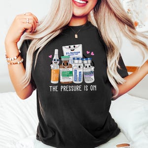 Puede incluir: Camiseta negra con un gr&aacute;fico de viales m&eacute;dicos de dibujos animados y una bolsa de soluci&oacute;n de dextrose. El texto "The Pressure Is On" est&aacute; impreso debajo del gr&aacute;fico.