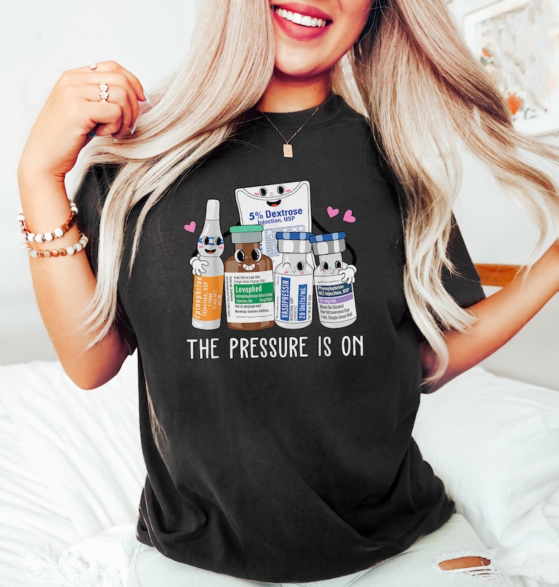 Puede incluir: Camiseta negra con un gr&aacute;fico de viales m&eacute;dicos de dibujos animados y una bolsa de soluci&oacute;n de dextrose. El texto "The Pressure Is On" est&aacute; impreso debajo del gr&aacute;fico.