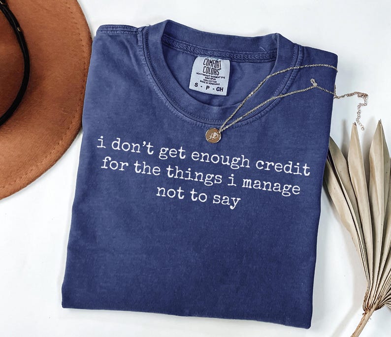 Puede incluir: Una camiseta Comfort Colors azul marino con el texto "i don't get enough credit for the things i manage not to say" en blanco. Tambi&eacute;n se ven un collar dorado y un sombrero marr&oacute;n.