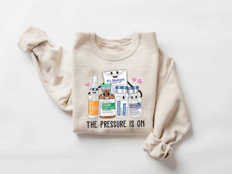 Puede incluir: Una sudadera beige con un gr&aacute;fico de suministros m&eacute;dicos de dibujos animados, que incluye una bolsa de soluci&oacute;n de dextrosa, un vial de levotiroxina y dos viales de insulina. El gr&aacute;fico incluye el texto "The Pressure Is On".