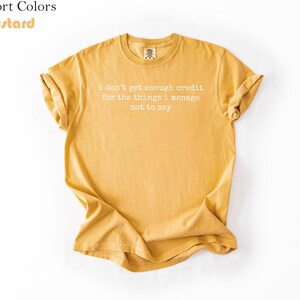 Puede incluir: Camiseta de color mostaza con el texto blanco "I don't get enough credit for the things I manage not to say". La camiseta es de cuello redondo y tiene mangas enrolladas.