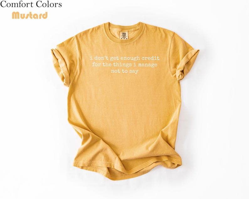 Puede incluir: Camiseta de color mostaza con el texto blanco "I don't get enough credit for the things I manage not to say". La camiseta es de cuello redondo y tiene mangas enrolladas.