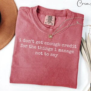 Puede incluir: Una camiseta de color carmes&iacute; con el texto blanco "i don't get enough credit for the things i manage not to say". Un collar dorado con un colgante est&aacute; sobre la camiseta. La camiseta est&aacute; doblada sobre una superficie blanca.