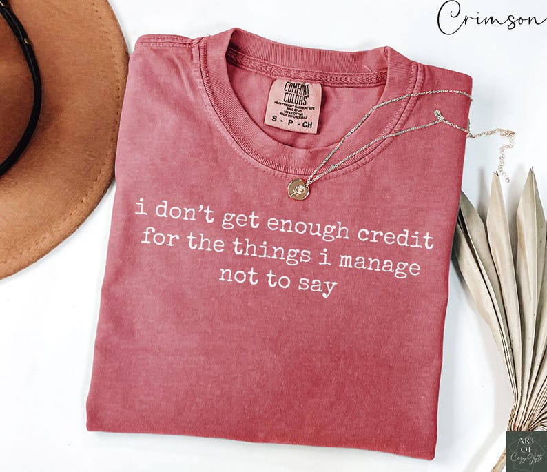 Puede incluir: Una camiseta de color carmes&iacute; con el texto blanco "i don't get enough credit for the things i manage not to say". Un collar dorado con un colgante est&aacute; sobre la camiseta. La camiseta est&aacute; doblada sobre una superficie blanca.