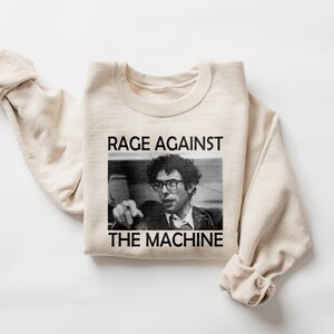 May include: A beige sweatshirt with the text "RAGE AGAINST THE MACHINE" printed on the front. The text is in black and there is a black and white photo of a man wearing glasses below the text.
