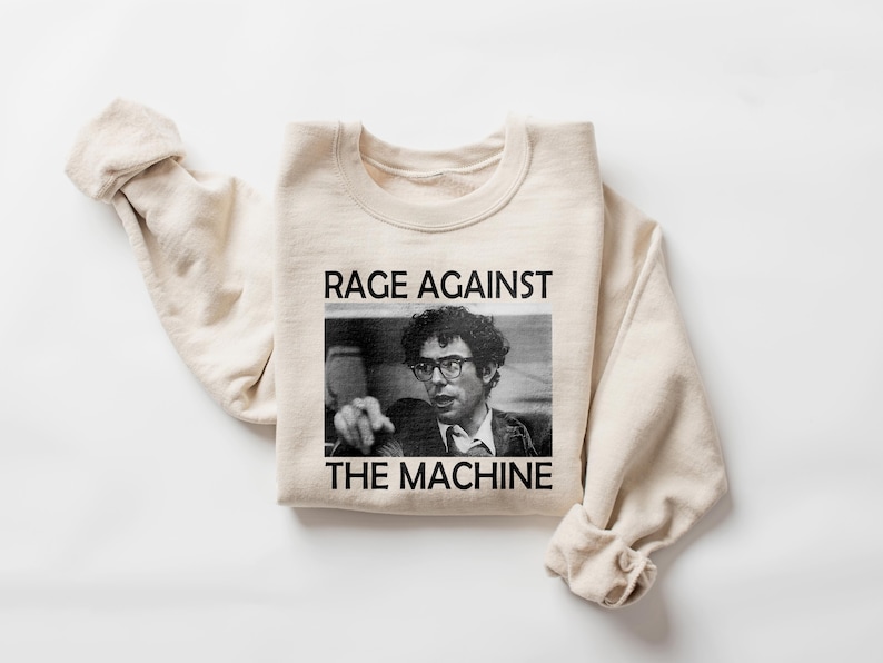 May include: A beige sweatshirt with the text "RAGE AGAINST THE MACHINE" printed on the front. The text is in black and there is a black and white photo of a man wearing glasses below the text.