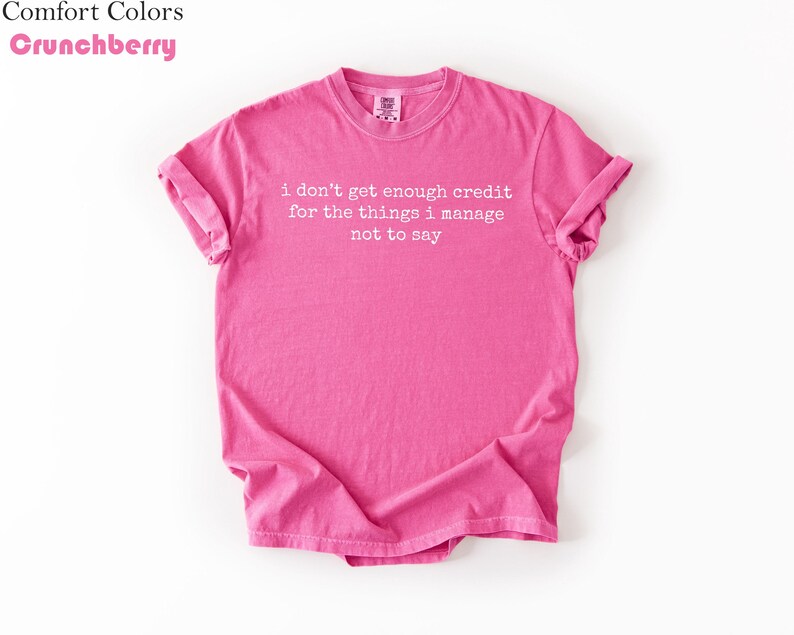 Puede incluir: Una camiseta rosa brillante con las mangas enrolladas. La camiseta presenta el texto "i don't get enough credit for the things i manage not to say" en blanco. La camiseta es de la marca Comfort Colors en color Crunchberry.