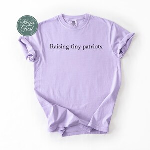 May include: A light purple t-shirt with the text "Raising tiny patriots." The shirt has short sleeves and a crew neck. A green and white sticker that says "Ships Fast" is in the upper left corner.
