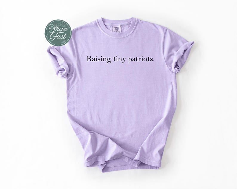 May include: A light purple t-shirt with the text "Raising tiny patriots." The shirt has short sleeves and a crew neck. A green and white sticker that says "Ships Fast" is in the upper left corner.