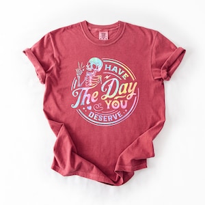 May include: A red t-shirt with a rainbow holographic graphic of a skeleton with the text "Have the Day You Deserve".
