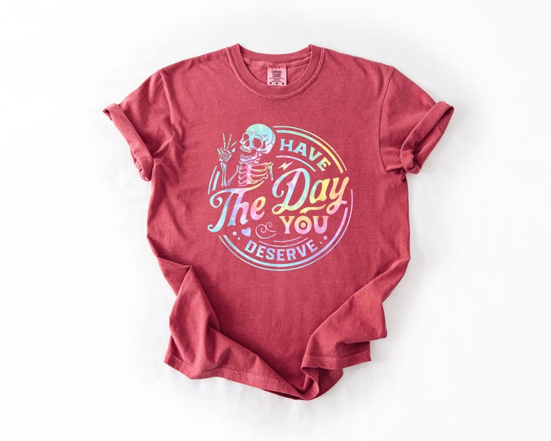 May include: A red t-shirt with a rainbow holographic graphic of a skeleton with the text "Have the Day You Deserve".