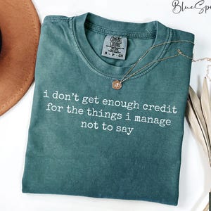 Puede incluir: Una camiseta Comfort Colors verde azulado con el texto "i don't get enough credit for the things i manage not to say" en blanco. Un collar dorado con un colgante est&aacute; sobre la camiseta.