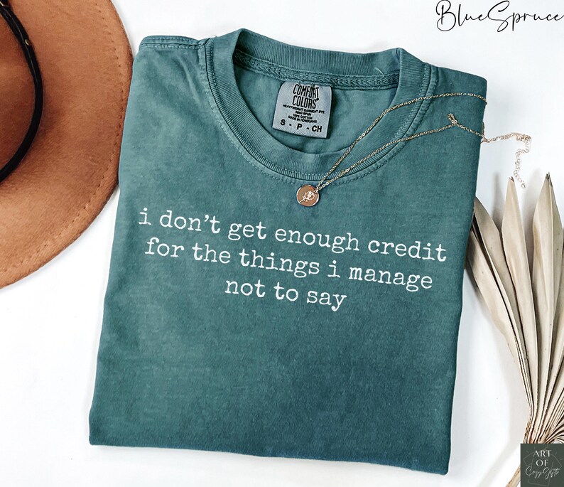 Puede incluir: Una camiseta Comfort Colors verde azulado con el texto "i don't get enough credit for the things i manage not to say" en blanco. Un collar dorado con un colgante est&aacute; sobre la camiseta.