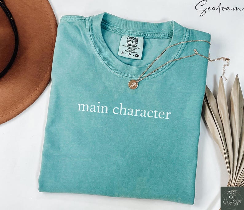 May include: Seafoam green t-shirt with the text "main character" in white lettering. The shirt is made by Comfort Colors and has a tag with the size and brand name.
