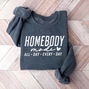 Puede incluir: Una sudadera gris oscuro con el texto "Homebody mode" en letras blancas y un peque&ntilde;o coraz&oacute;n blanco. El texto "All day every day" tambi&eacute;n est&aacute; en letras blancas debajo del texto principal.