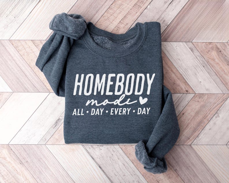 Puede incluir: Una sudadera gris oscuro con el texto "Homebody mode" en letras blancas y un peque&ntilde;o coraz&oacute;n blanco. El texto "All day every day" tambi&eacute;n est&aacute; en letras blancas debajo del texto principal.