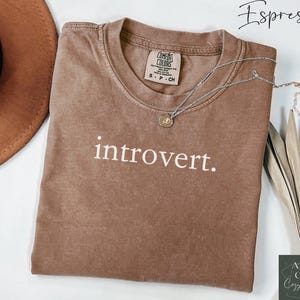 May include: A brown t-shirt with the text "introvert." printed in white. The shirt is laid flat on a white surface with a brown hat and a silver necklace.