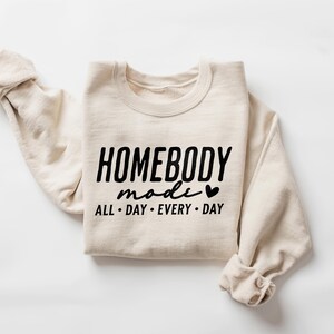 Puede incluir: Una sudadera beige con el texto "HOMEBODY mode ALL DAY EVERY DAY" y un peque&ntilde;o gr&aacute;fico de coraz&oacute;n impreso en la parte delantera.