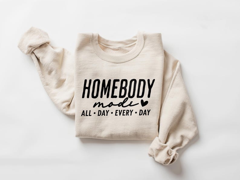 Puede incluir: Una sudadera beige con el texto "HOMEBODY mode ALL DAY EVERY DAY" y un peque&ntilde;o gr&aacute;fico de coraz&oacute;n impreso en la parte delantera.