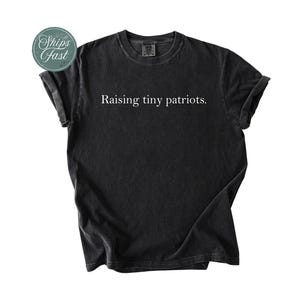 May include: Black t-shirt with the text "Raising tiny patriots." The shirt has a crew neck and short sleeves. A green and white sticker that says "Ships Fast" is in the upper left corner.