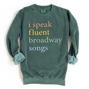May include: A dark green sweatshirt with the text "i speak fluent broadway songs" printed in rainbow colors.