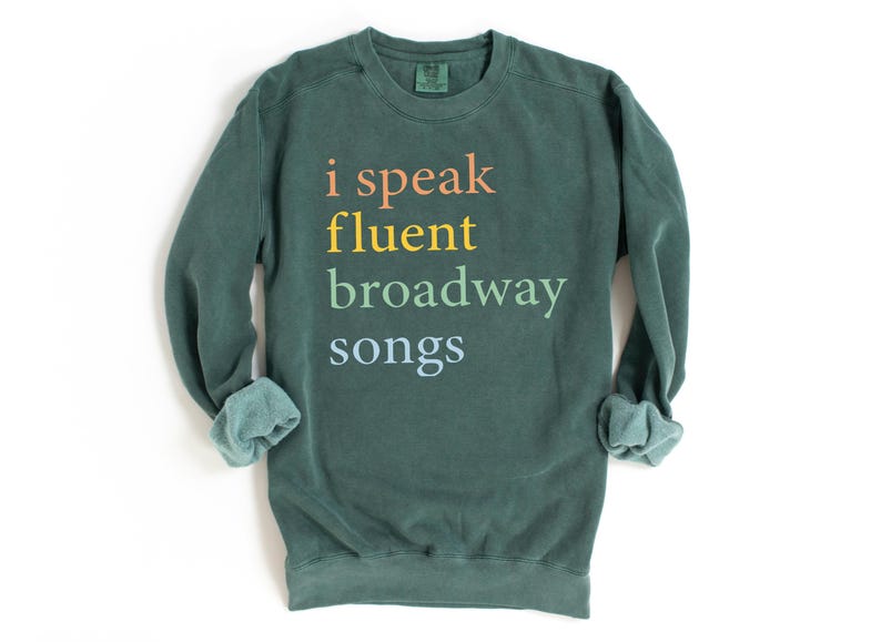 May include: A dark green sweatshirt with the text "i speak fluent broadway songs" printed in rainbow colors.