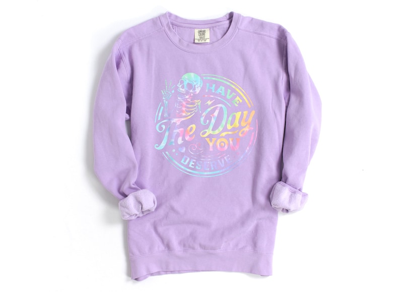 May include: A lavender long-sleeved sweatshirt with a rainbow tie-dye design featuring a skull and the text "Have the Day You Deserve".