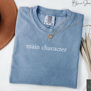 May include: A blue jean t-shirt with the text "main character" in white lettering. The shirt is folded and laid flat on a white surface.