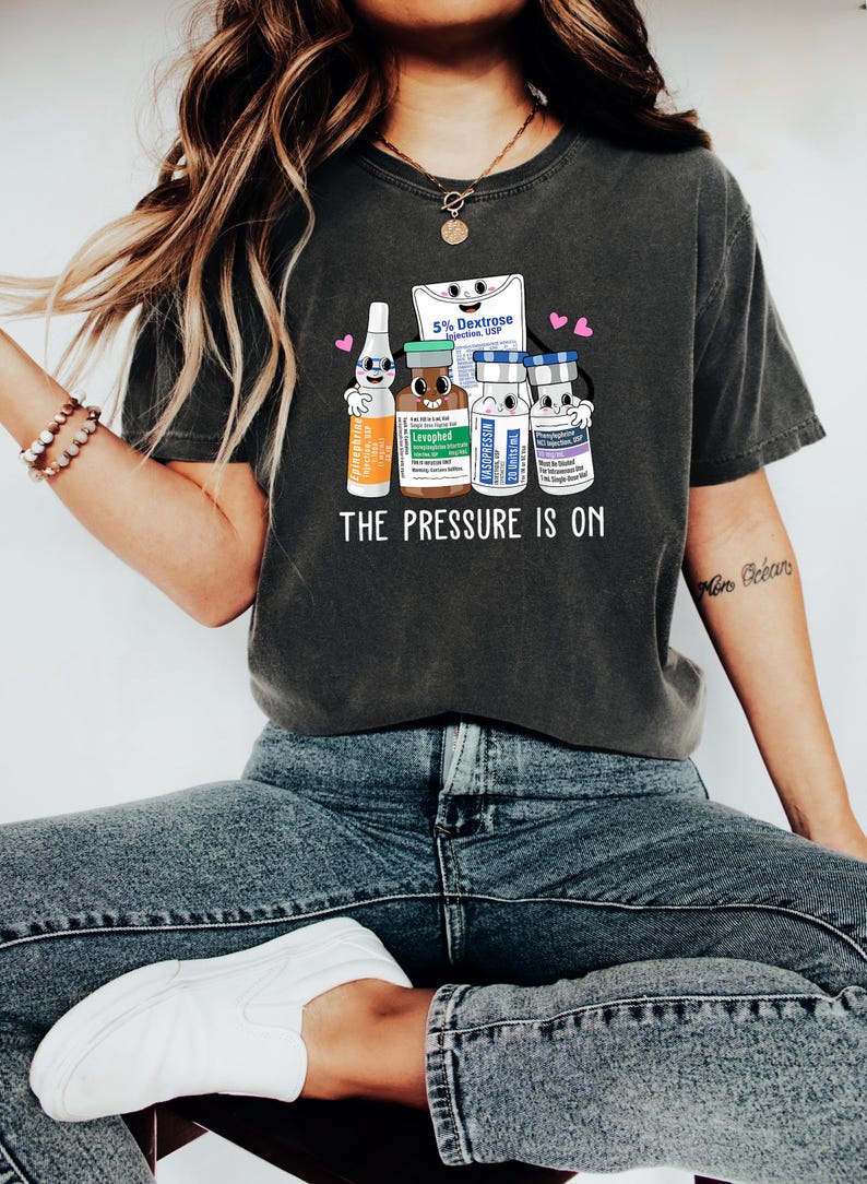 Puede incluir: Una camiseta gris oscuro con un gr&aacute;fico de suministros m&eacute;dicos de dibujos animados y el texto "THE PRESSURE IS ON".