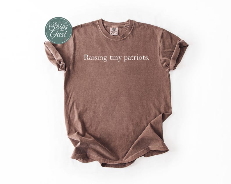 May include: A brown t-shirt with the text "Raising tiny patriots" in white. The shirt has a crew neck and short sleeves. The image also includes a green and white sticker that says "Ships fast".